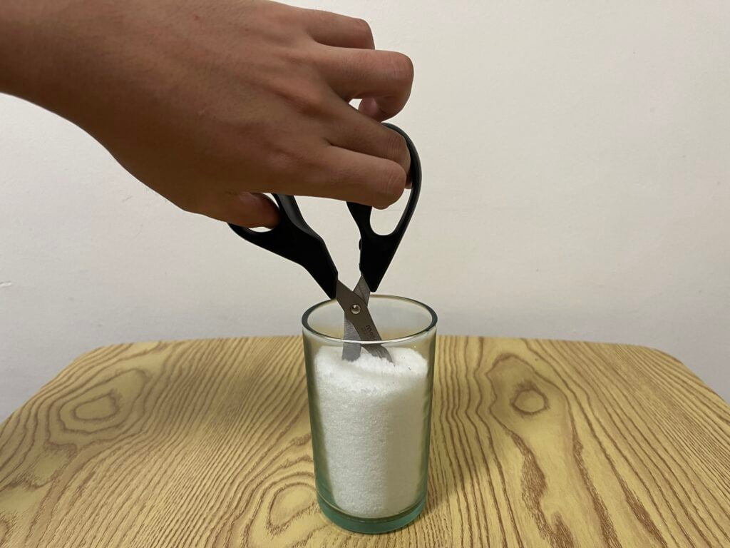 A hand uses scissors to cut into a clear glass filled to the top with white granulated substance, placed on a wooden surface.