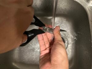 A person is washing a pair of black-handled scissors under running water in a stainless steel sink.