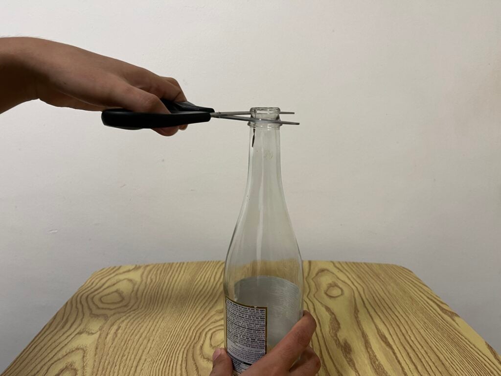 A person uses scissors to open the metal cap of a glass bottle on a wooden table.