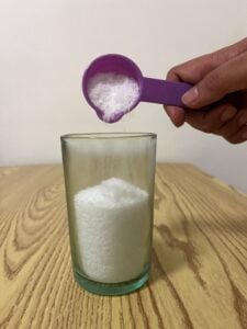 A hand pours white granules from a purple scoop into a glass filled with similar white granules on a wooden table.