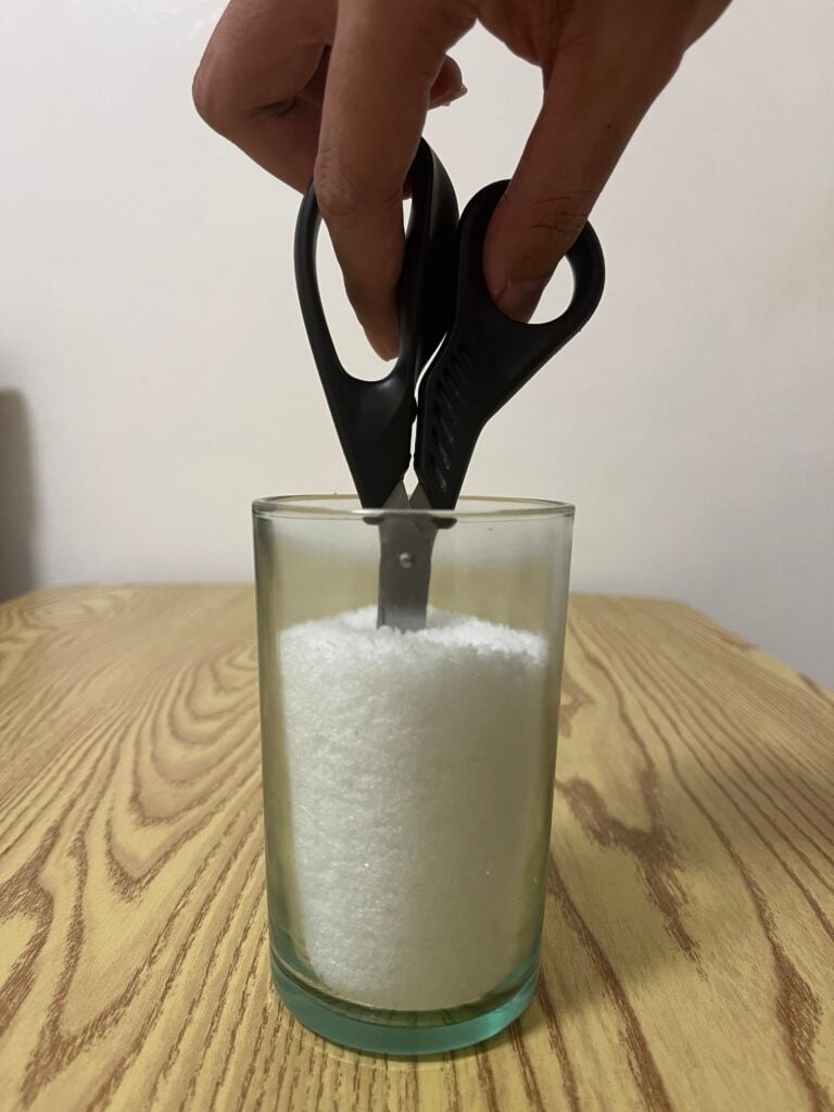 A hand holds scissors, blade down, inserted into a glass filled with white granules, placed on a wooden surface.