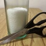 A glass filled with white granules sits on a wood table, with a pair of black-handled scissors resting against it.