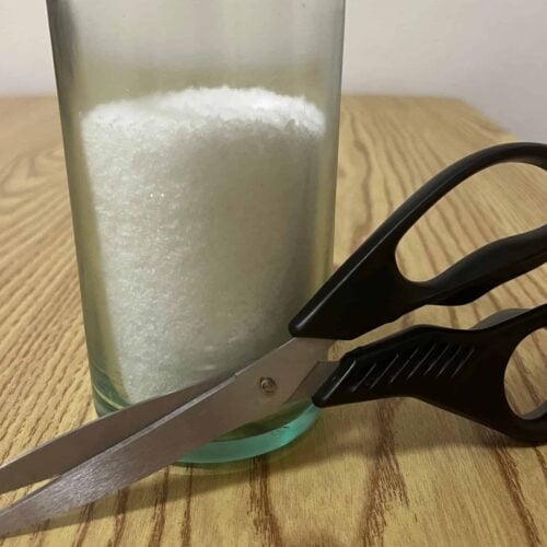 A glass filled with white granules sits on a wood table, with a pair of black-handled scissors resting against it.