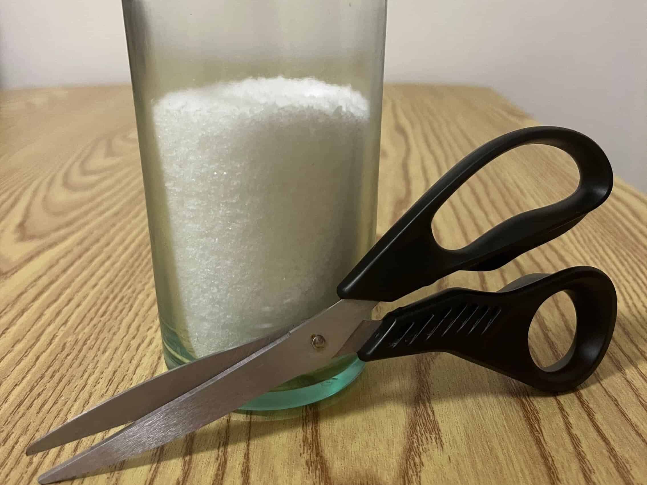 A glass filled with white granules sits on a wood table, with a pair of black-handled scissors resting against it.
