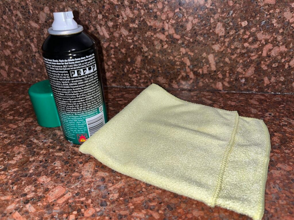 A can of spray cleaner with a green cap and a yellow microfiber cloth are placed on a brown granite countertop.