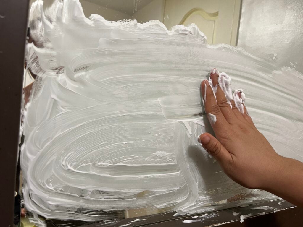 A hand spreads white shaving cream on a bathroom mirror, covering most of the surface with thick, swirling layers.