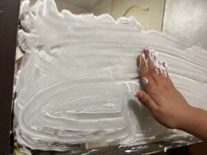 A hand spreads white shaving cream on a bathroom mirror, covering most of the surface with thick, swirling layers.