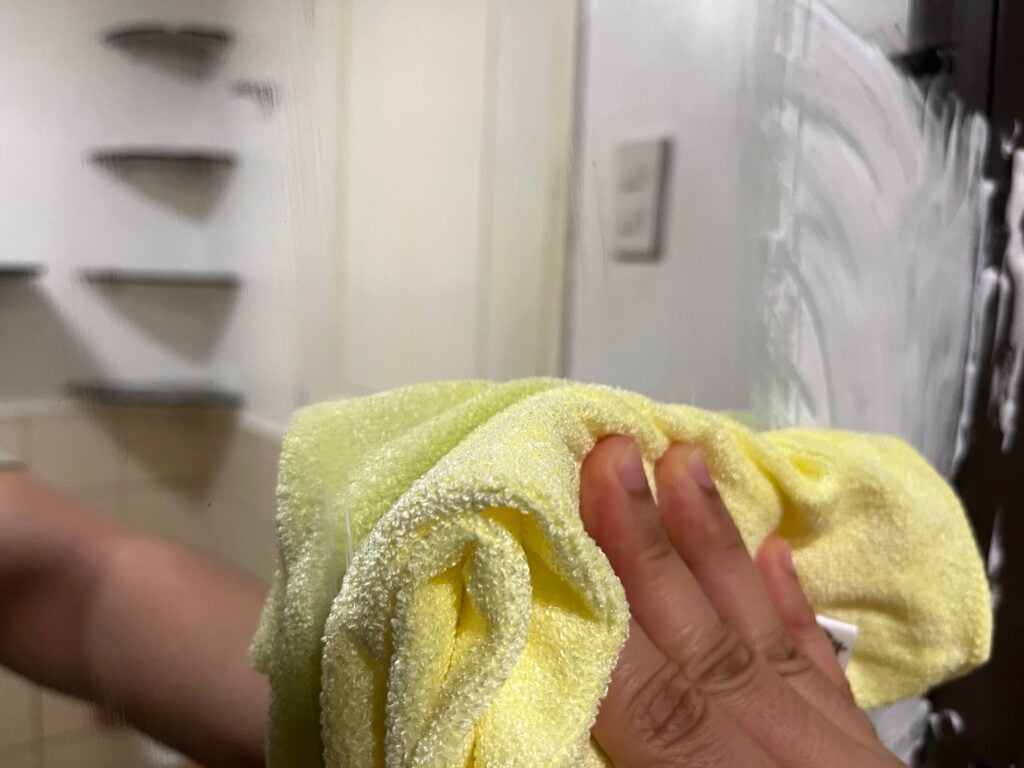 A hand holding a yellow microfiber cloth wipes a mirror, removing soap or cleaner residue.