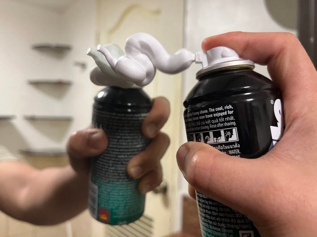 A hand sprays shaving foam onto a mirror, creating a swirl of foam between the can nozzle and its reflection.