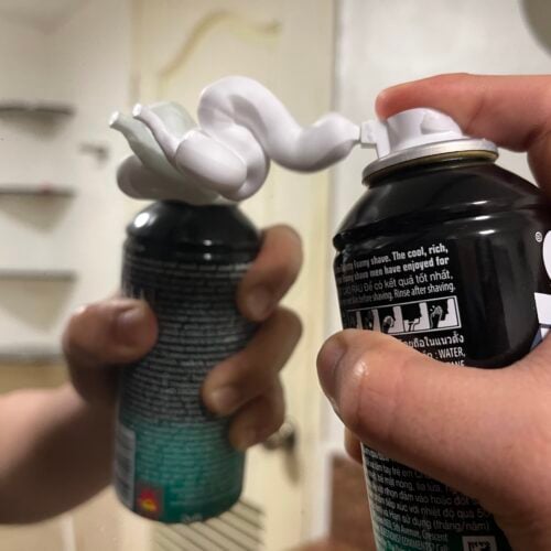 A hand sprays shaving foam onto a mirror, creating a swirl of foam between the can nozzle and its reflection.