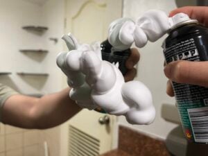 A person sprays thick white foam from a can onto a mirror, creating twisted, cloud-like shapes.