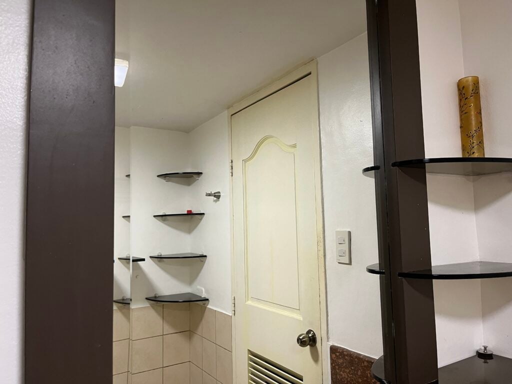 A beige door with a silver knob is seen in a bathroom with white walls, brown shelves, and beige tile. A yellow canister is on the right shelf.