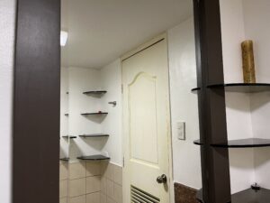 A beige door with a silver knob is seen in a bathroom with white walls, brown shelves, and beige tile. A yellow canister is on the right shelf.