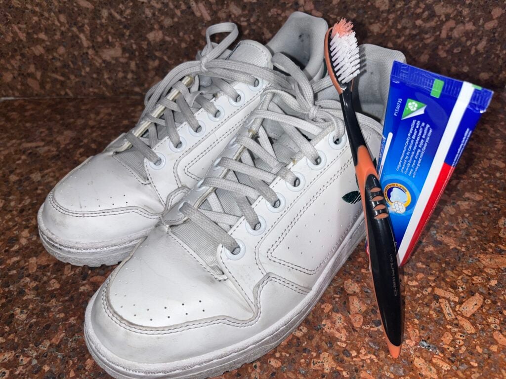 A pair of white sneakers, a black and orange toothbrush with toothpaste, and a blue packet of cleaning wipes on a brown textured surface.