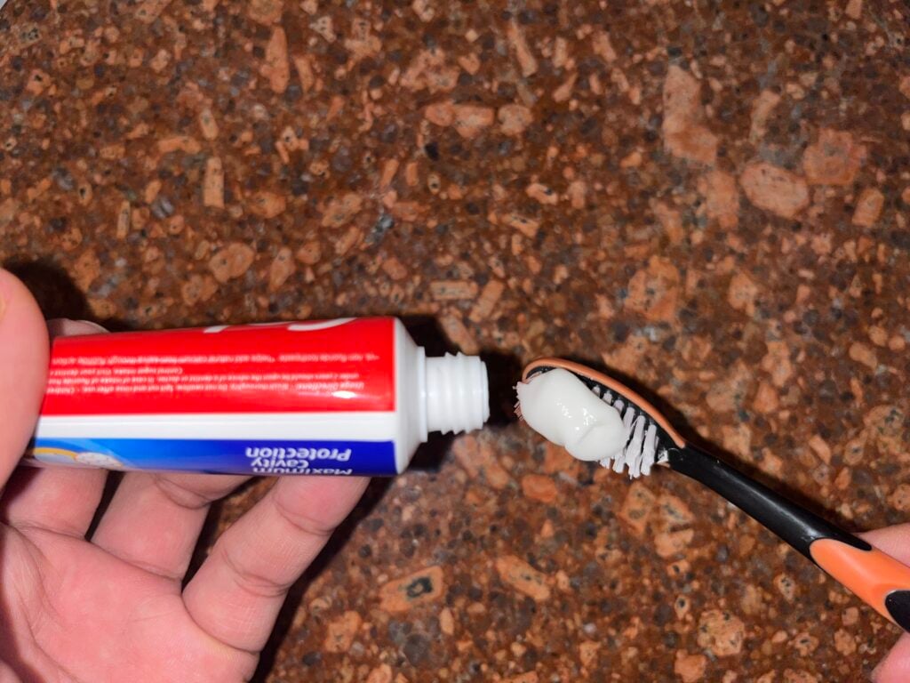 A hand squeezes toothpaste from a red and white tube onto a black toothbrush over a brown speckled countertop.
