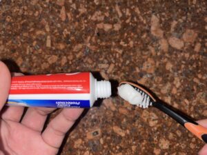 A hand squeezes toothpaste from a red and white tube onto a black toothbrush over a brown speckled countertop.