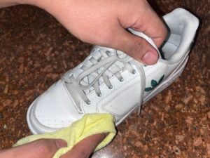 A person cleans a white sneaker with a yellow cloth on a brown speckled surface.