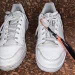 Two white sneakers on a brown surface; the right shoe has an orange and black toothbrush placed across its laces.