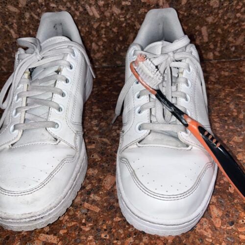 Two white sneakers on a brown surface; the right shoe has an orange and black toothbrush placed across its laces.