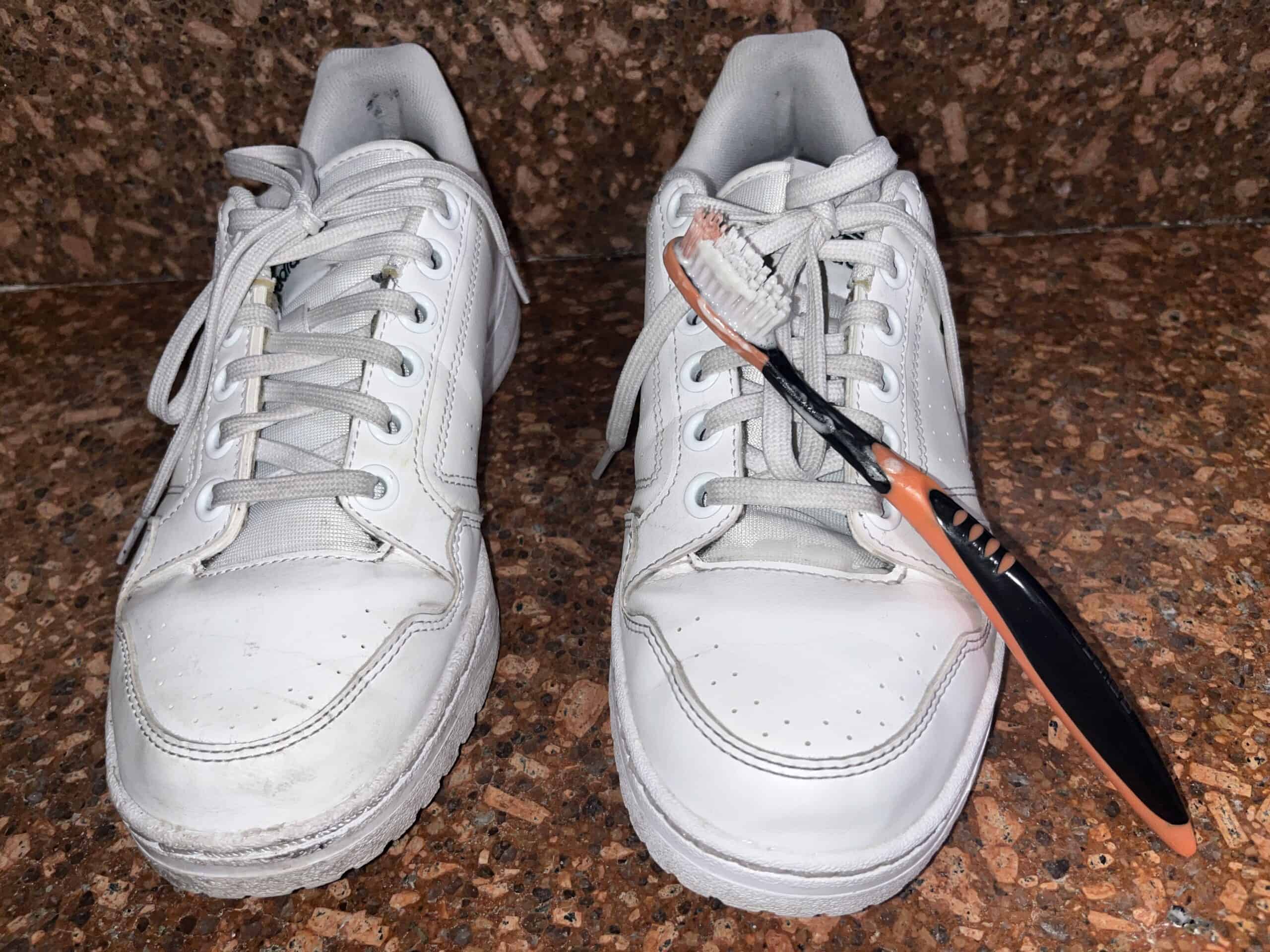 Two white sneakers on a brown surface; the right shoe has an orange and black toothbrush placed across its laces.