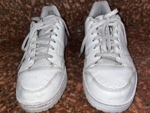 A pair of white sneakers with laces, placed side by side on a speckled brown surface. The shoes show slight signs of wear, including minor creases and smudges.