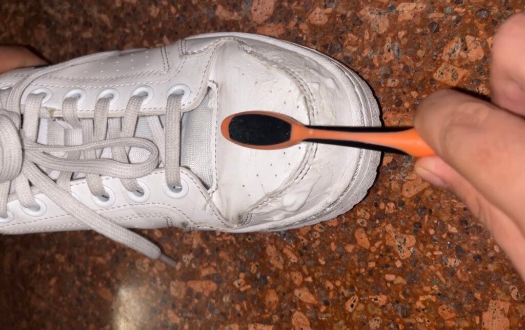 A white sneaker is being cleaned with a black and orange toothbrush on a brown speckled surface.
