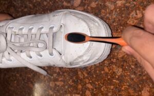 A white sneaker is being cleaned with a black and orange toothbrush on a brown speckled surface.