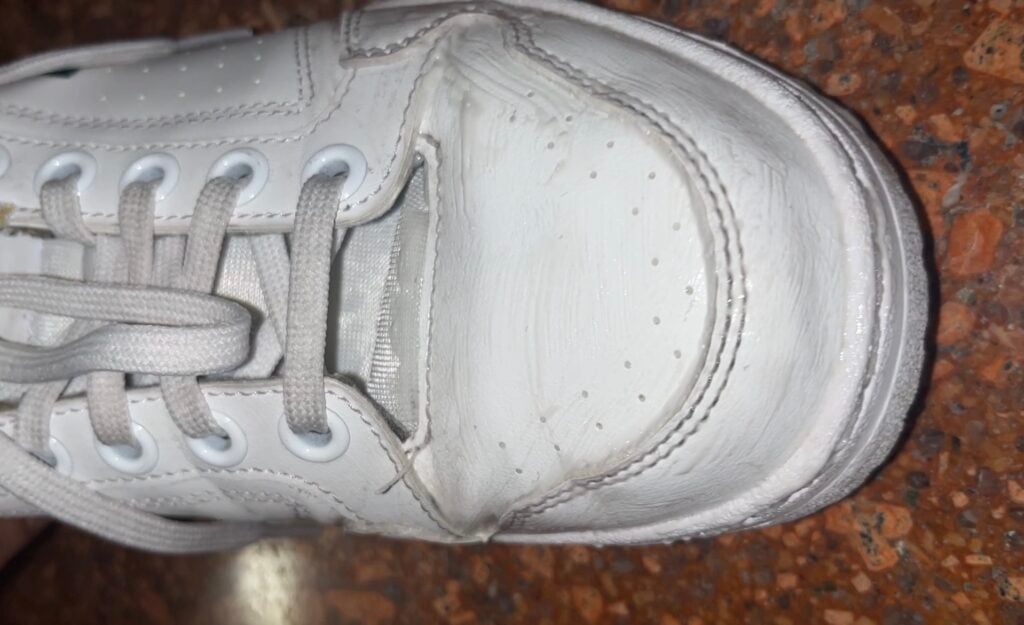 White sneaker with visible damage at the front, showing a large tear in the upper material, on a speckled brown floor.