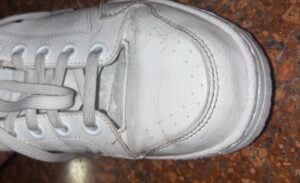 White sneaker with visible damage at the front, showing a large tear in the upper material, on a speckled brown floor.