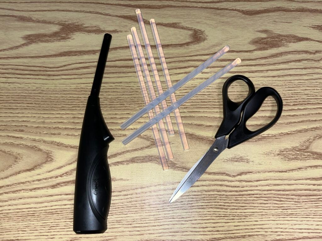 A black lighter, five plastic straws, and a pair of black scissors are arranged on a light wood-grain surface.