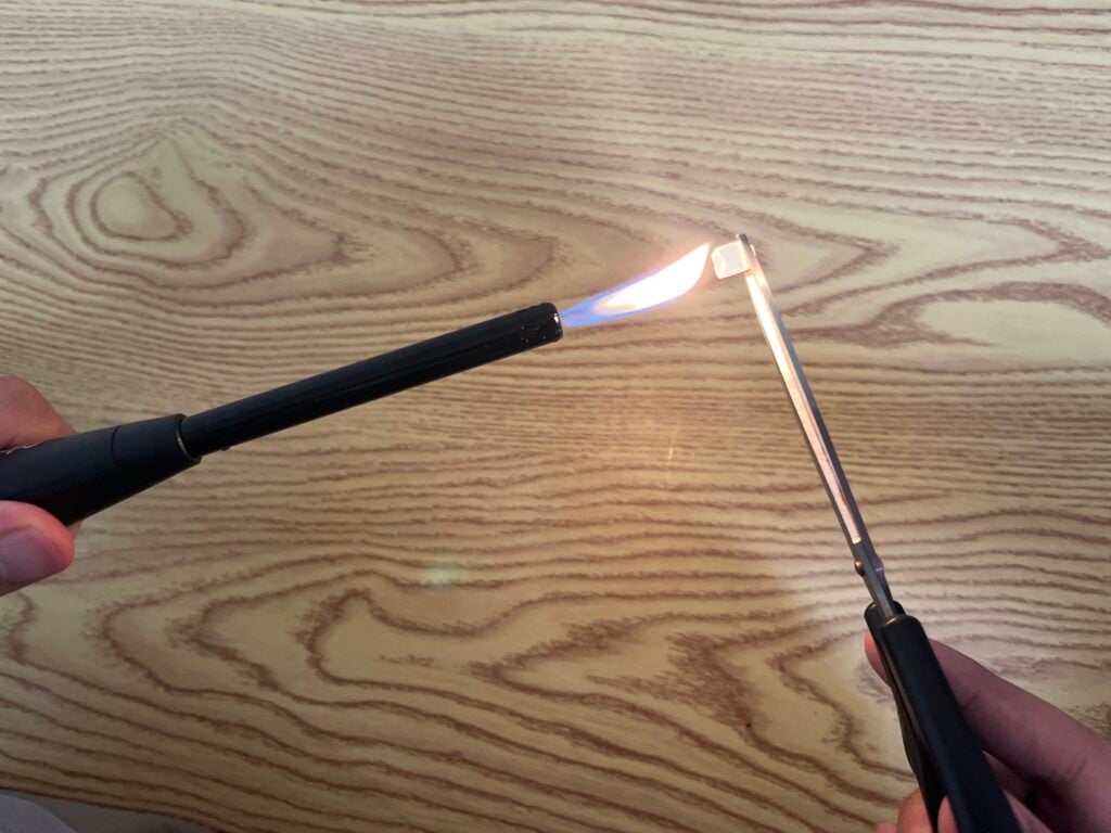 A person lights a candle lighter with a lit match, holding the match and lighter over a wood-patterned surface.