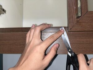 A person uses scissors to scrape an object from the top edge of a wooden cabinet door while holding it steady with their other hand.
