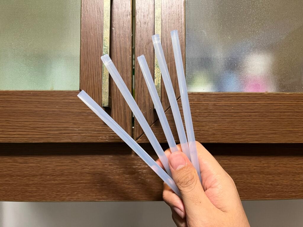 A hand holds five translucent plastic straws in front of a wooden-framed frosted glass window.