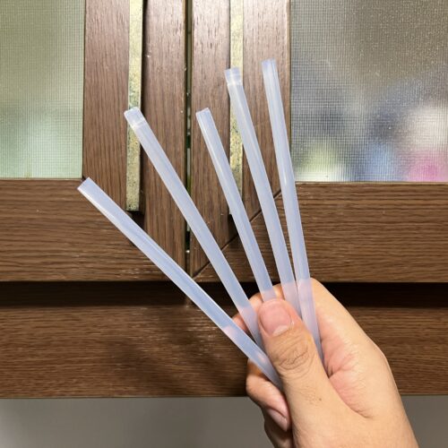 A hand holds five translucent plastic straws in front of a wooden-framed frosted glass window.