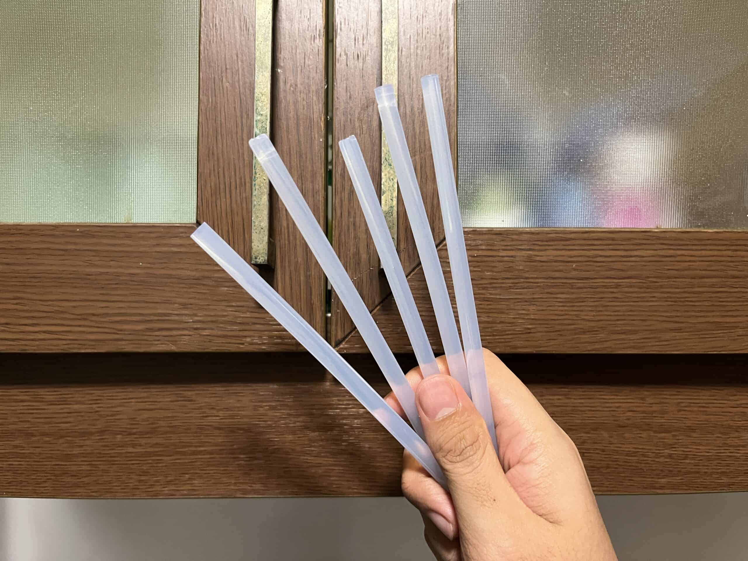 A hand holds five translucent plastic straws in front of a wooden-framed frosted glass window.