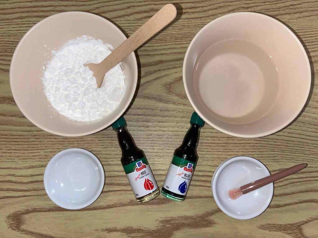 Two bowls, one with white powder and one with clear liquid, sit on a wooden surface, along with red and blue food coloring and two small empty bowls with wooden stirrers.