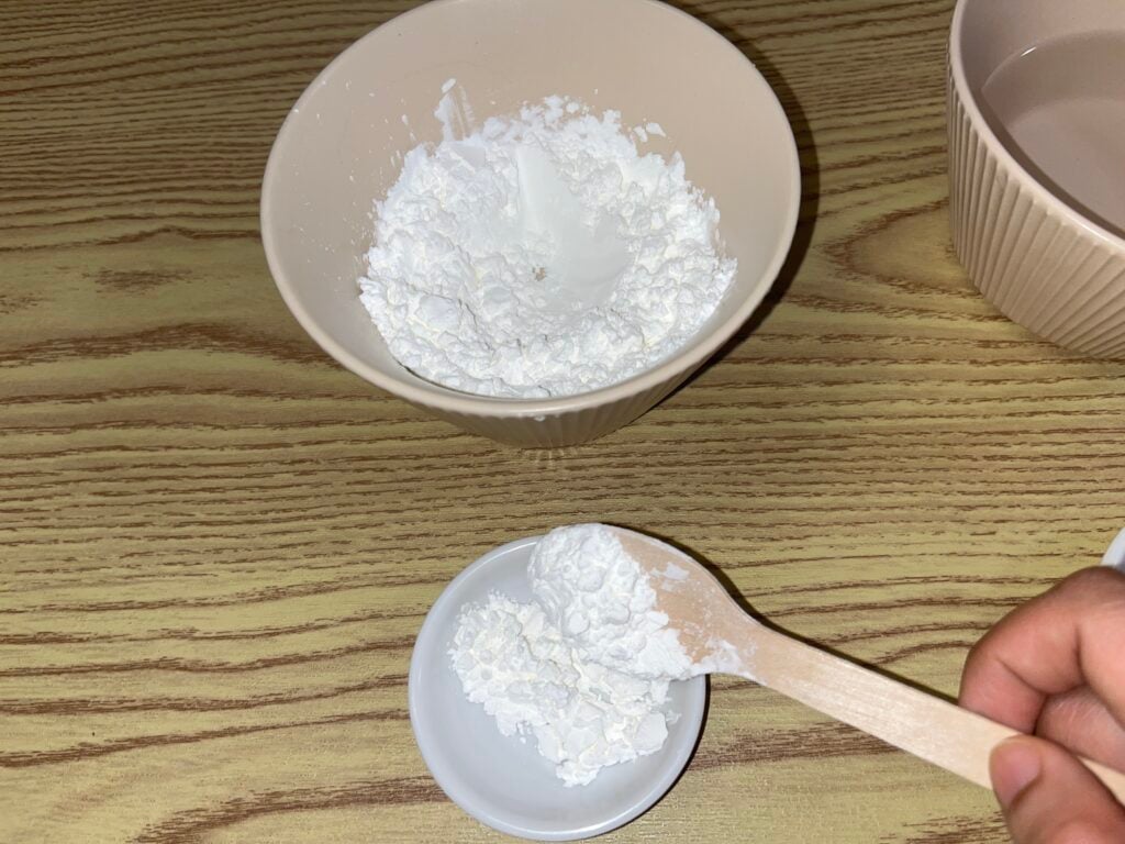 A hand spoons white powder, possibly cornstarch or flour, from a large bowl into a small dish on a wooden surface.