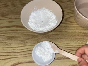A hand spoons white powder, possibly cornstarch or flour, from a large bowl into a small dish on a wooden surface.