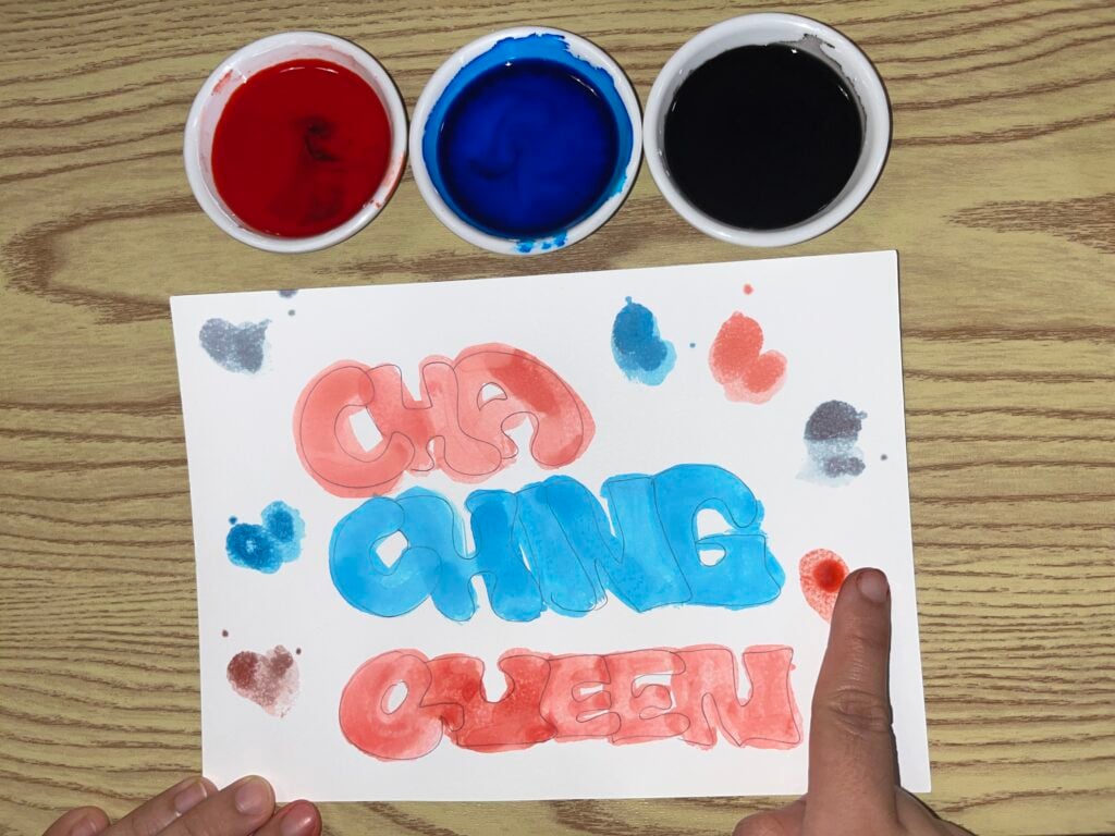 A hand points to a paper with “CHA CHING QUEEN” painted in red and blue bubble letters, surrounded by paint splatters. Three bowls of red, blue, and black paint are above the paper on a wood surface.
