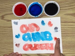 A hand points to a paper with “CHA CHING QUEEN” painted in red and blue bubble letters, surrounded by paint splatters. Three bowls of red, blue, and black paint are above the paper on a wood surface.