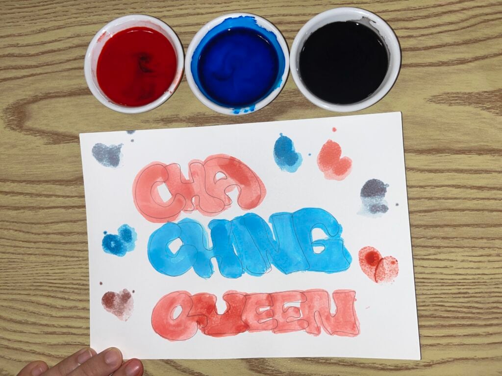 A hand holds a paper on a wood table. “CHA CHING QUEEN” is painted in red and blue, with three small bowls of red, blue, and black paint above the paper.