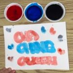 A hand holds a paper on a wood table. “CHA CHING QUEEN” is painted in red and blue, with three small bowls of red, blue, and black paint above the paper.