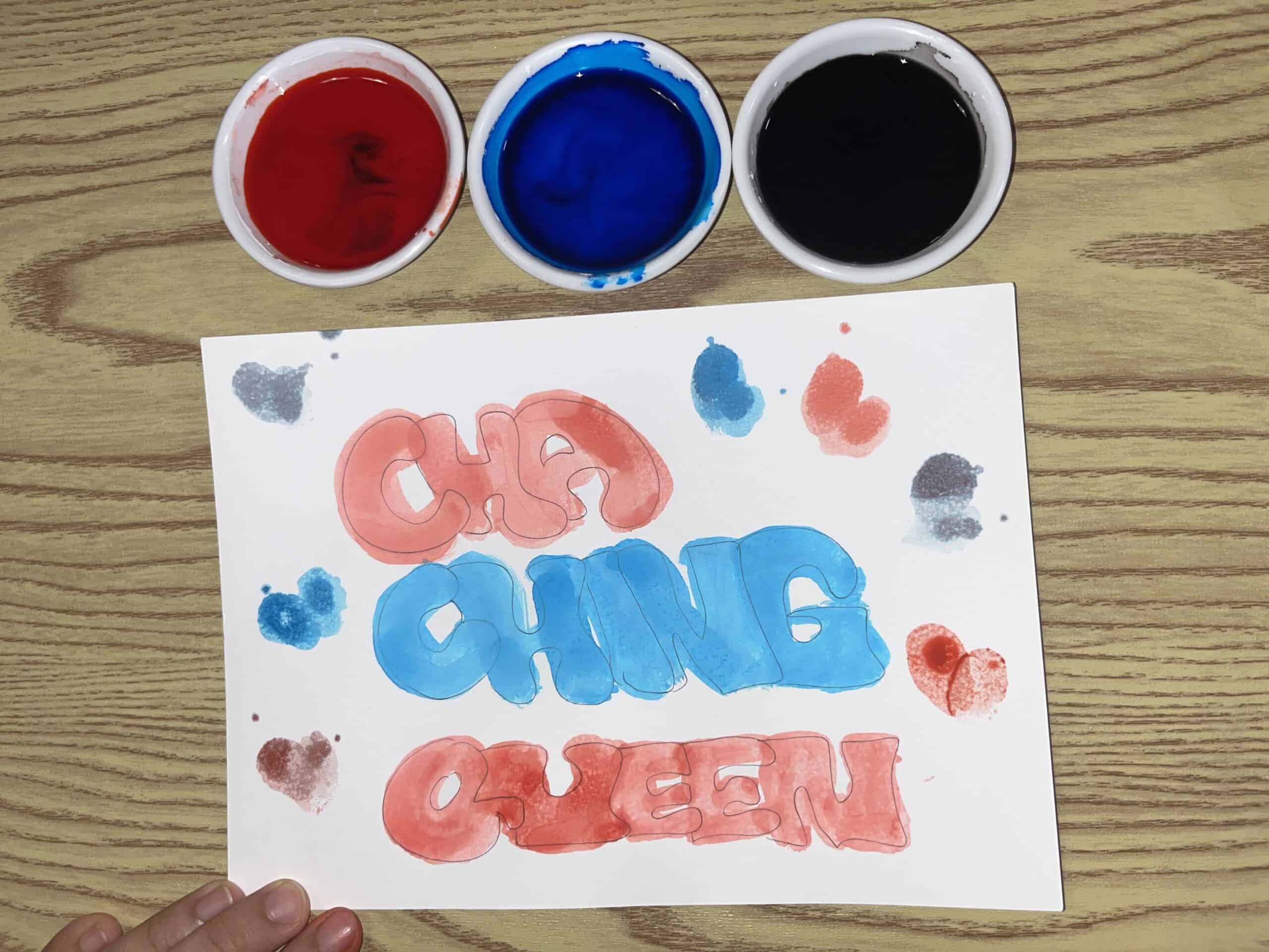A hand holds a paper on a wood table. “CHA CHING QUEEN” is painted in red and blue, with three small bowls of red, blue, and black paint above the paper.