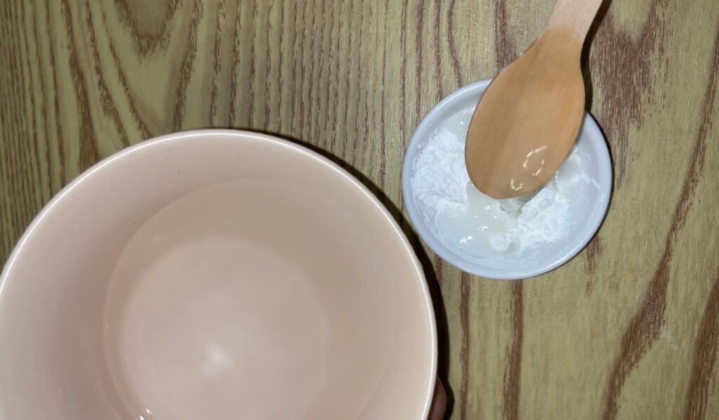 A large beige bowl sits next to a small white bowl containing white powder and a wooden spoon on a wooden surface.