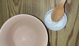 A large beige bowl sits next to a small white bowl containing white powder and a wooden spoon on a wooden surface.