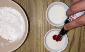 A hand adds red food coloring to a small bowl of white liquid next to another similar bowl and a plate of white powder on a wooden surface.