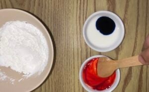 A bowl of white powder, a small dish of white liquid with blue dye, and a dish of red liquid being stirred with a wooden spoon are arranged on a wooden surface.