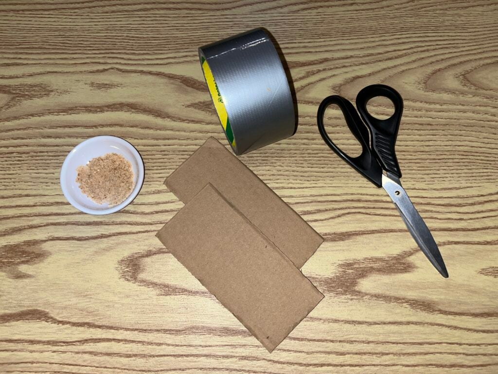 A small bowl of light-colored powder, a roll of duct tape, two pieces of cardboard, and a pair of scissors on a wood-grain surface.