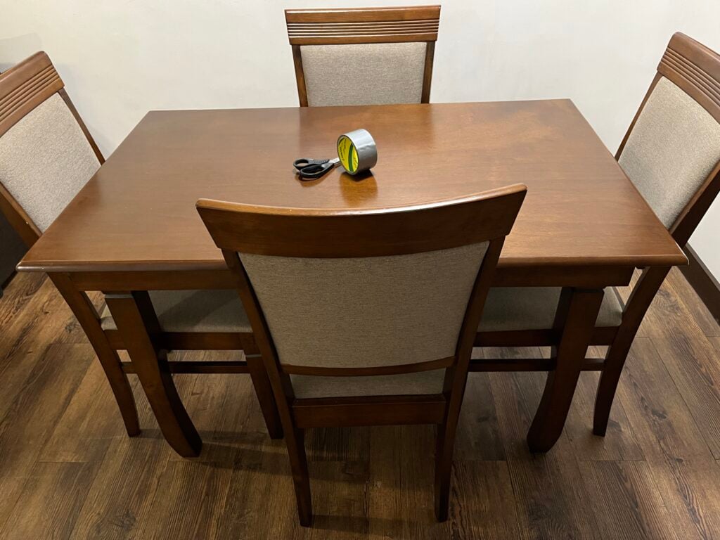 A wooden dining table with four chairs and a roll of duct tape with scissors in the center.
