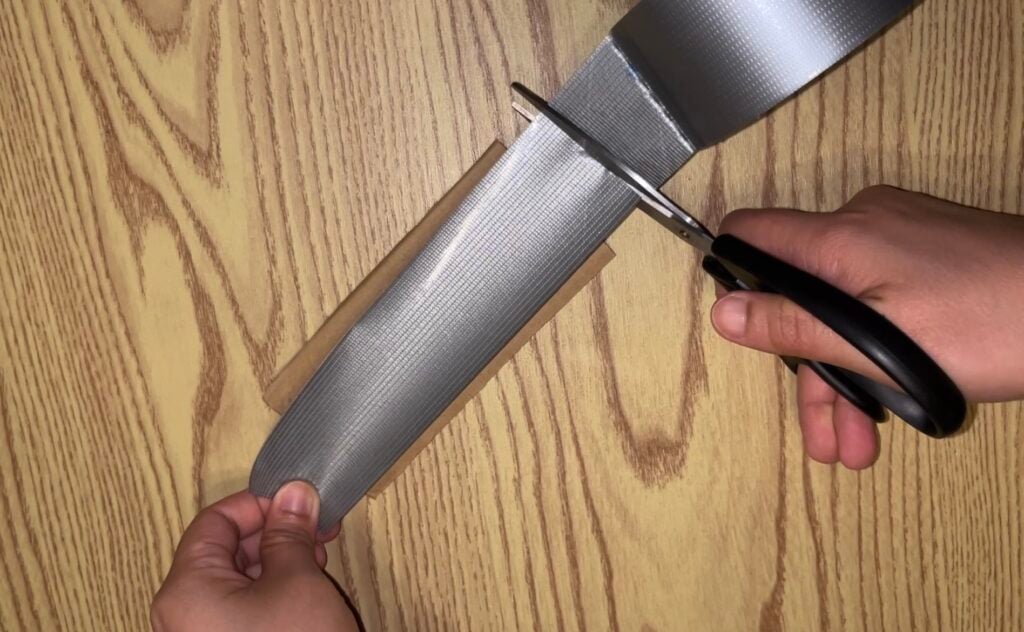 Hands using scissors to cut a strip of gray duct tape on a wooden surface.